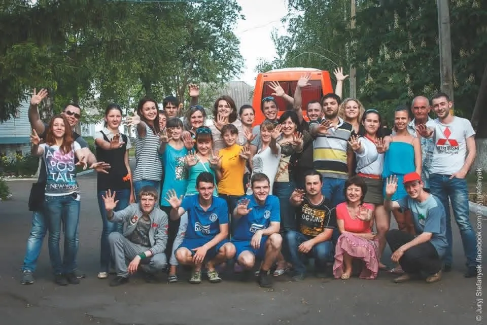 A large group of volunteers waving at the camera in front of an orange bus, trees around