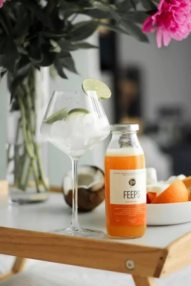 Bottle of citrus drink on a wooden tray beside a wine glass with ice and lime, peonies in the background