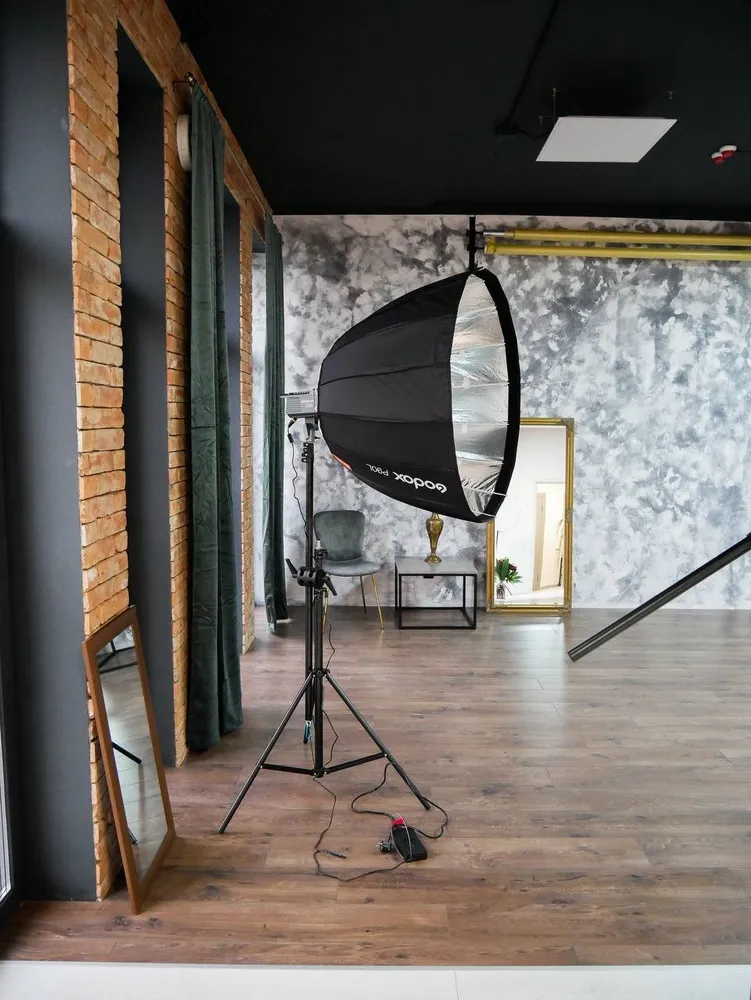 Photography studio interior: large parabolic softbox on a stand, textured gray backdrop, brick wall and green velvet curtains