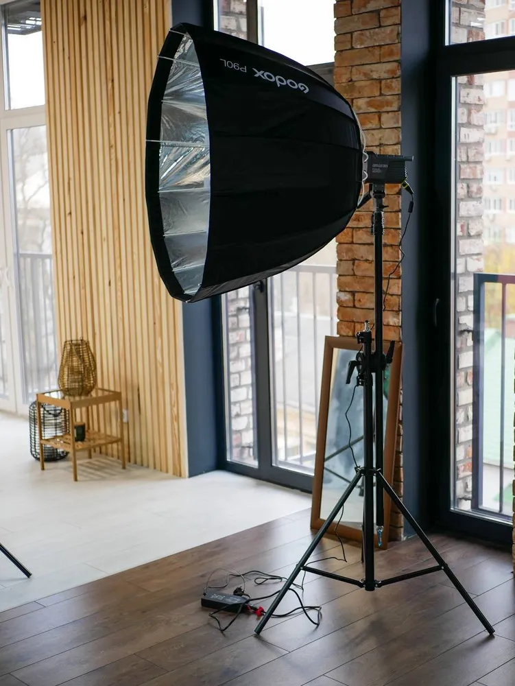 Studio corner by tall windows: parabolic softbox and LED light on stands, brick column, wooden slat wall