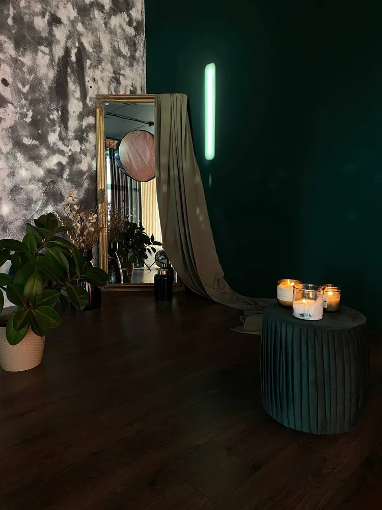 Dark styled studio corner: gold-framed mirror with draped fabric, candles on a green velvet pouf, plants