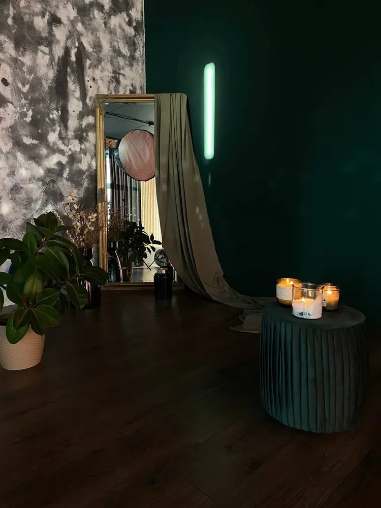 Dark styled studio corner: gold-framed mirror with draped fabric, candles on a green velvet pouf, plants